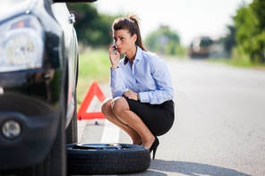 Picture of a woman on her phone on the side of the road needing to change a tire