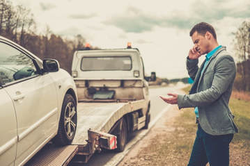 Picture of man on side of the road on his phone who is upset that he is getting his car towed